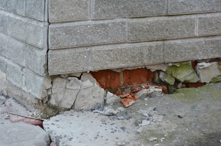 Crumbling foundation at the base of a block wall, with broken concrete and exposed red brick