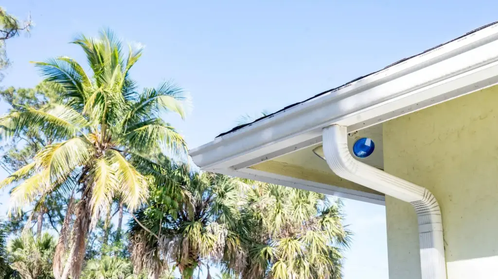 A white gutter and downspout on a yellow house, with palm trees against a blue sky