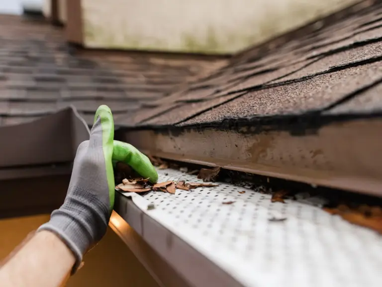 A gloved hand clearing leaves off a gutter guard at the edge of a shingle roof