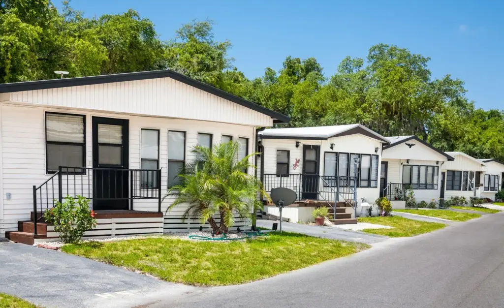A row of white manufactured homes with small porches and palm plantings along a paved street