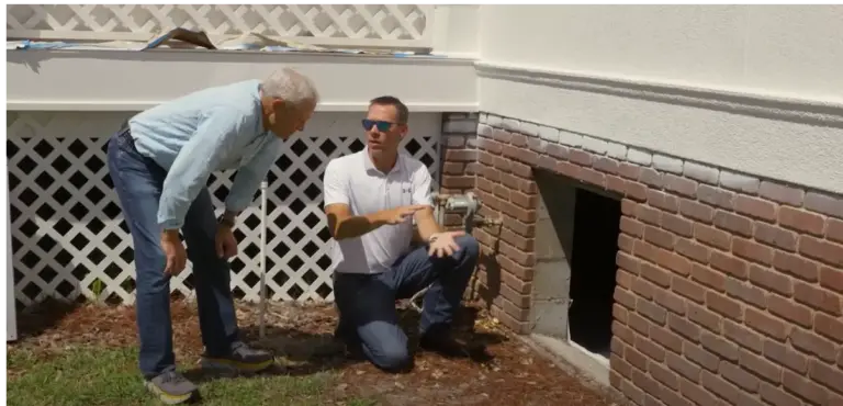 A technician kneels at a crawl space opening in a brick foundation wall, talking with a homeowner