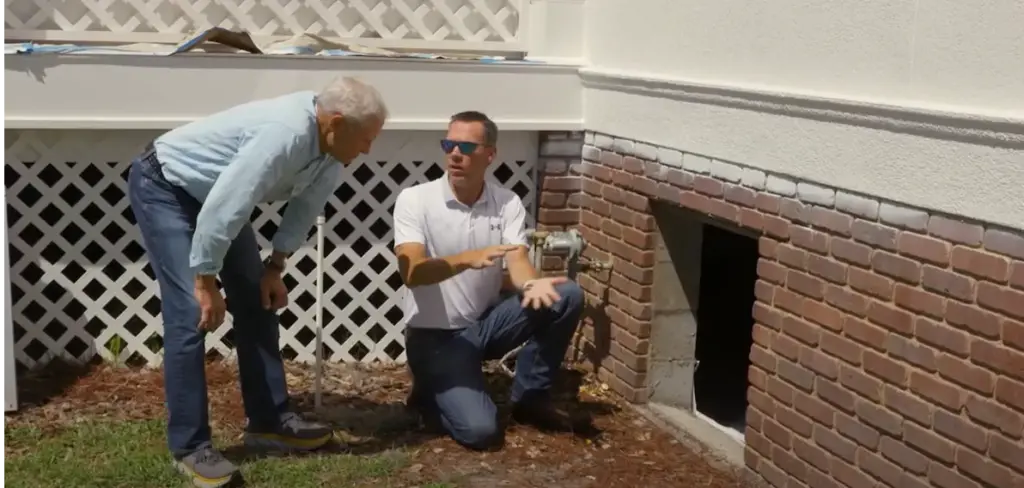 A Metro Foundation Solutions specialist kneels by a crawl space opening in a brick foundation, explaining what he found to a homeowner